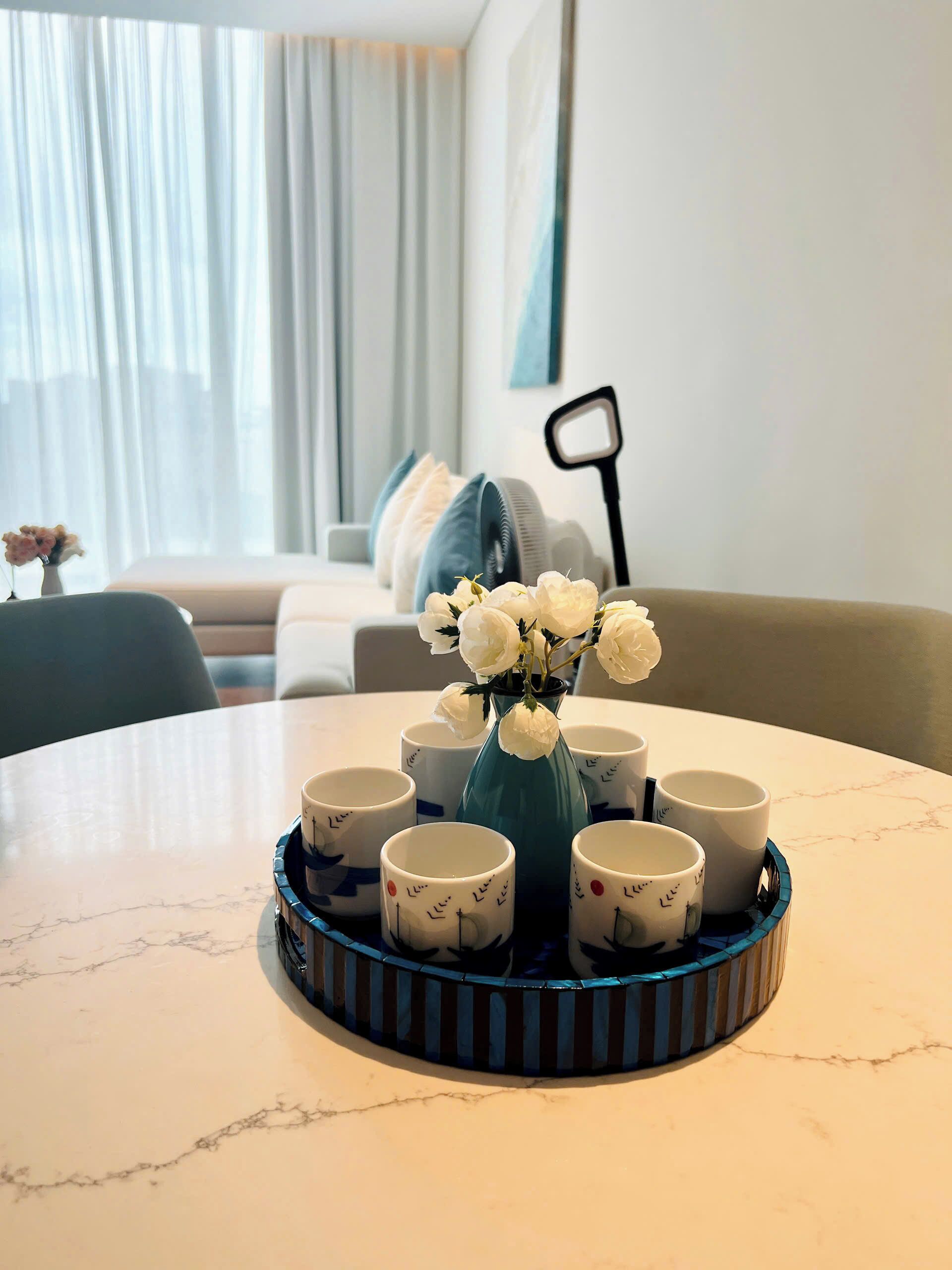 A round table with a vase of white flowers and a tray of white mugs with blue designs.