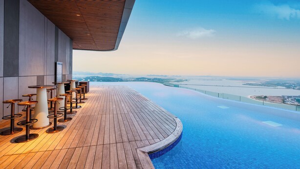 A rooftop infinity pool with a wooden deck and bar stools, overlooking a cityscape. - Alacarte Ha Long Bay Managed by Navis Global (Bai Chay)