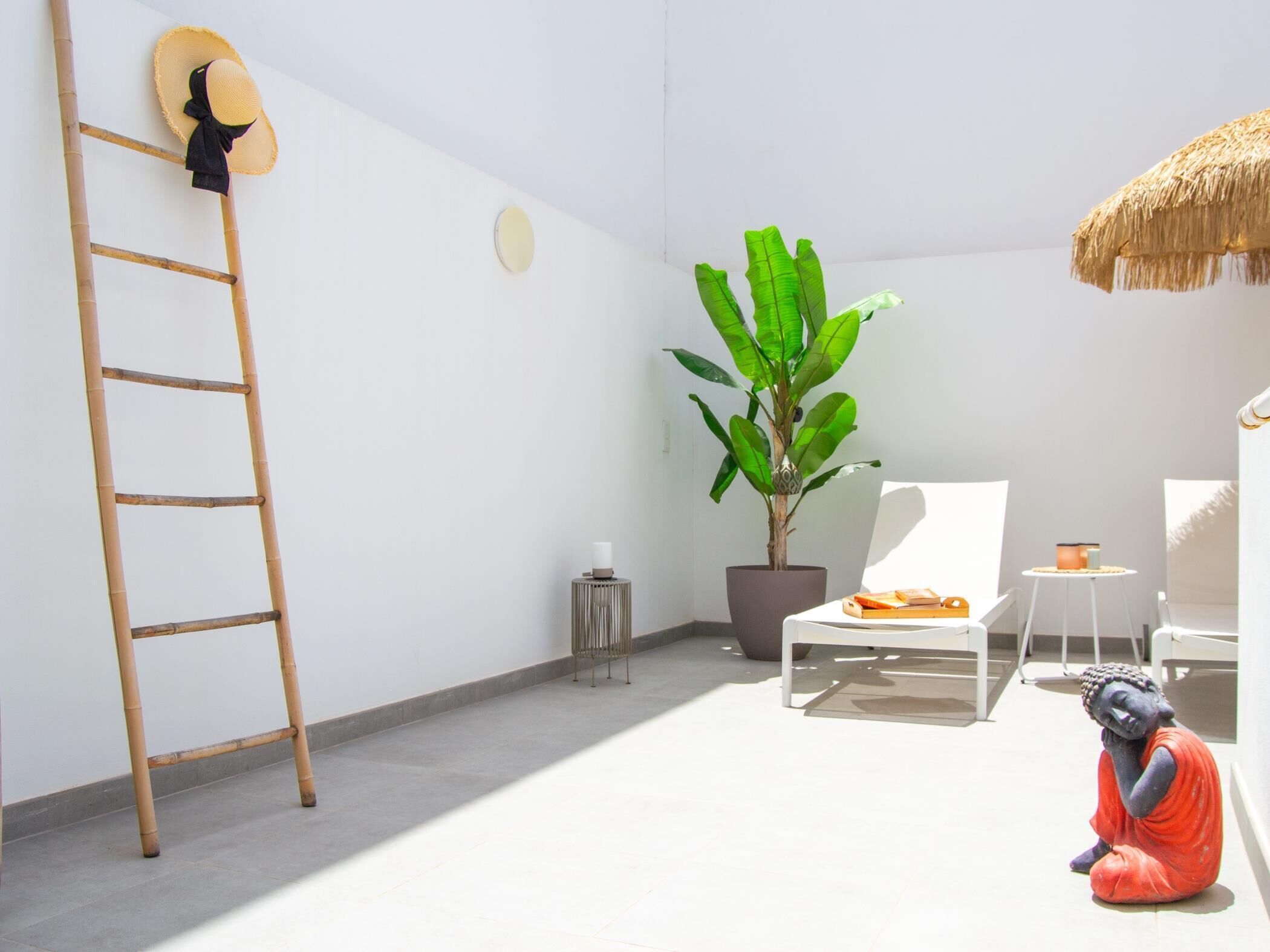 A minimalist room with a ladder, a large potted plant, a white chair, a small table, and a Buddha statue.
