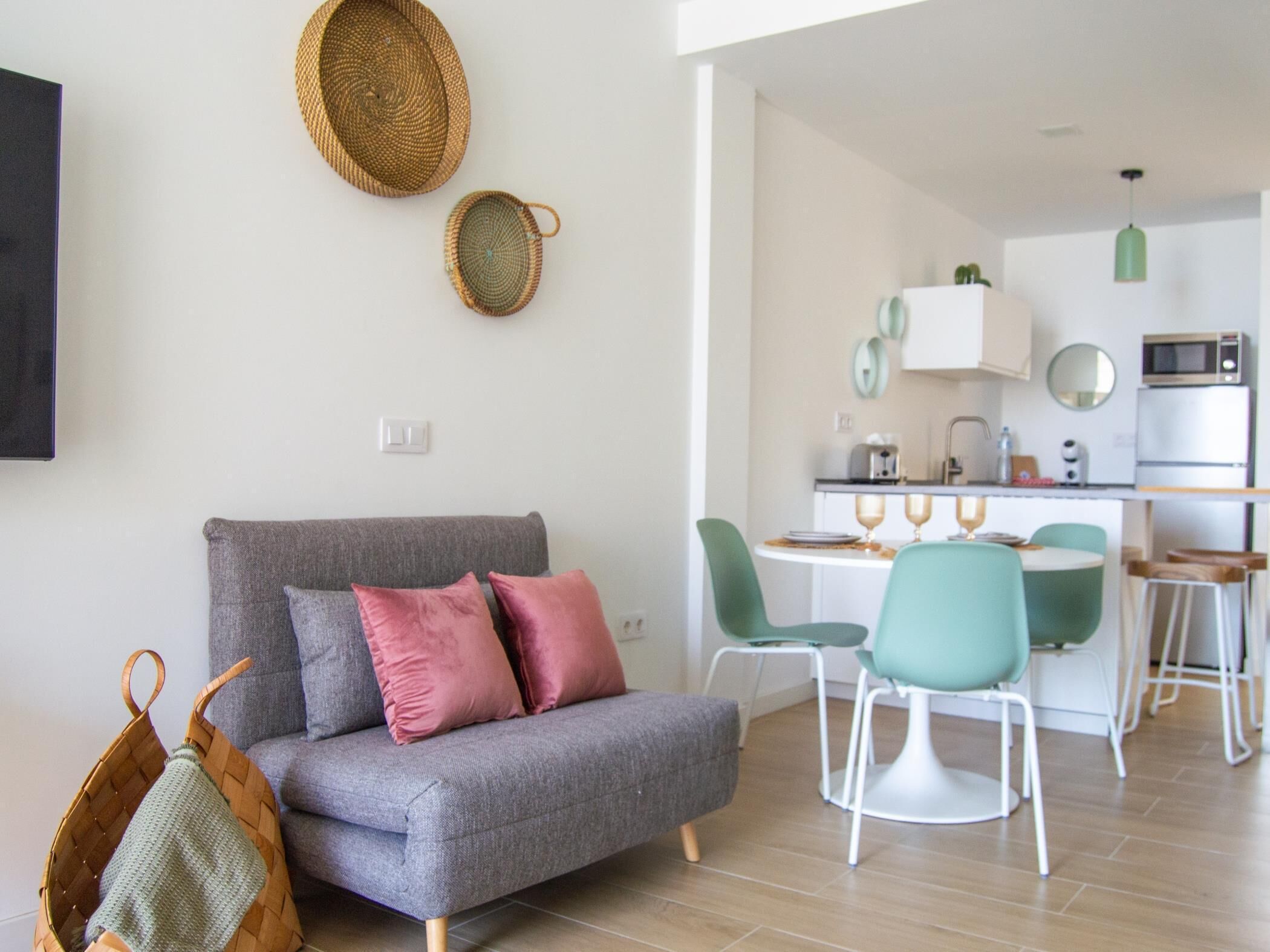 Ein modernes Wohnzimmer mit grauem Sofa, zwei Kissen und einem gewebten Korb. Es gibt einen Essbereich mit Tisch und Stühlen sowie eine Küche mit Mikrowelle und Backofen.