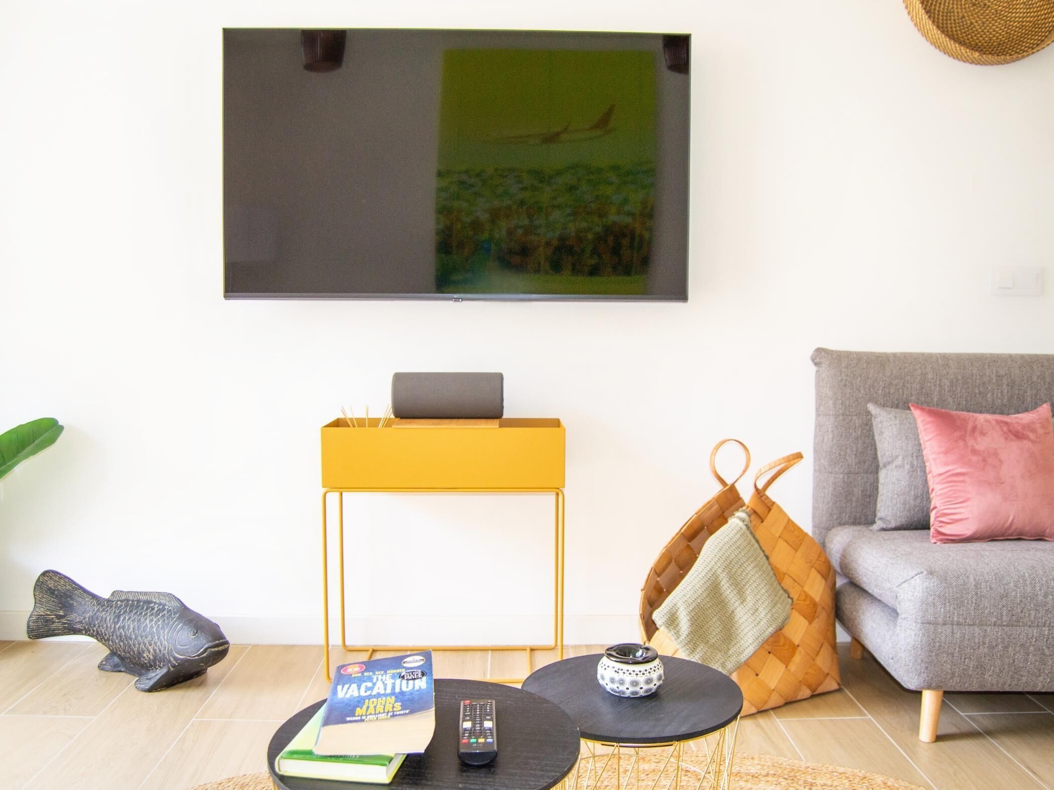 A living room with a sofa, a coffee table, a TV mounted on the wall, and a small side table with a book and remote control.