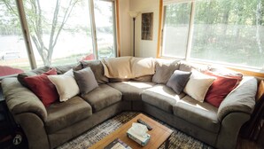 Un sofá seccional en forma de L con varios cojines, una mesa de centro con libros y un control remoto, y ventanas amplias que ofrecen vistas a árboles y un lago.