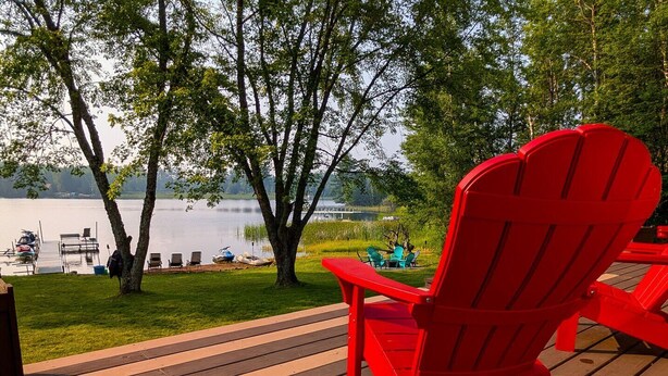 Una terraza con una silla Adirondack roja con vista a un lago, rodeada de árboles y barcos amarrados.