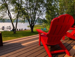Una terraza con una silla Adirondack roja con vista a un lago, rodeada de árboles y barcos amarrados.