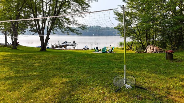 Un área con césped, una cancha de bádminton, un bote amarrado junto al agua y árboles al fondo.