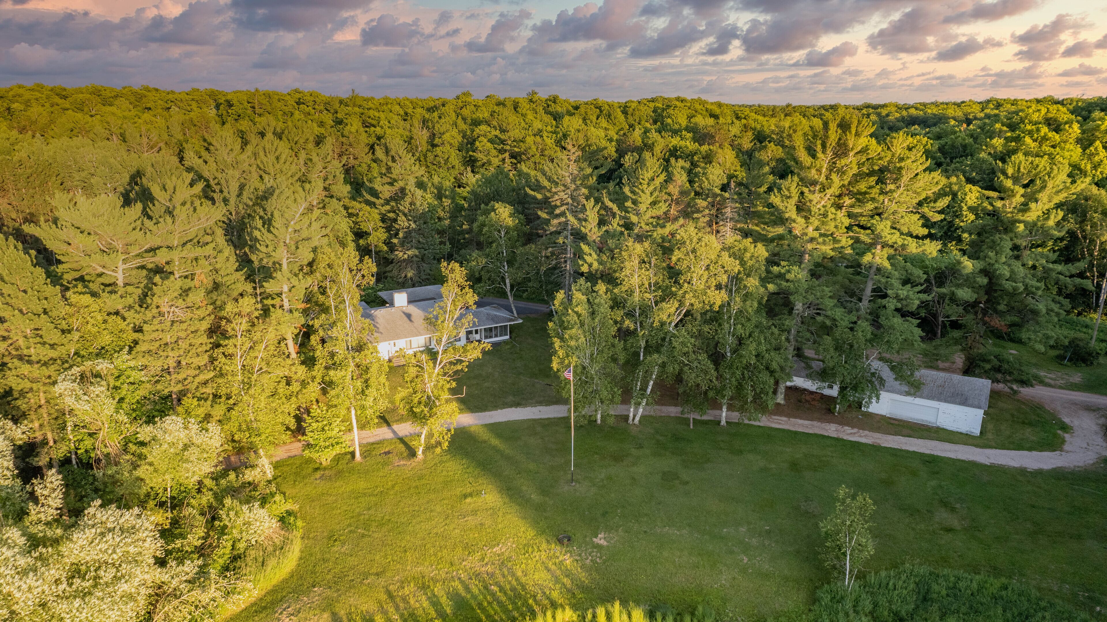 Une vue aérienne d’une maison entourée d’une forêt dense, avec une allée bien dégagée et un mât de drapeau.