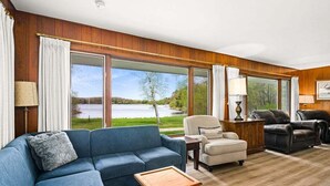 Un salon avec un canapé bleu, une petite table et deux chaises. De grandes fenêtres offrent une vue sur un lac et des arbres.