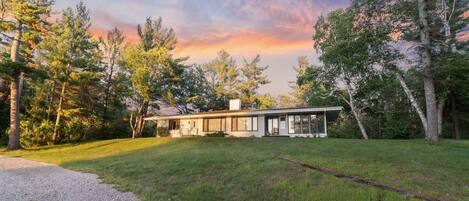 A modern house with a large front lawn, surrounded by trees and a clear sky.