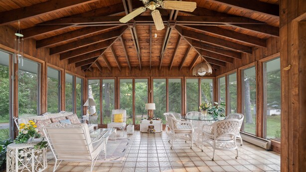 Une pièce spacieuse avec un plafond haut, de grandes fenêtres et un sol carrelé. On y trouve des chaises en osier blanc et une table en verre.