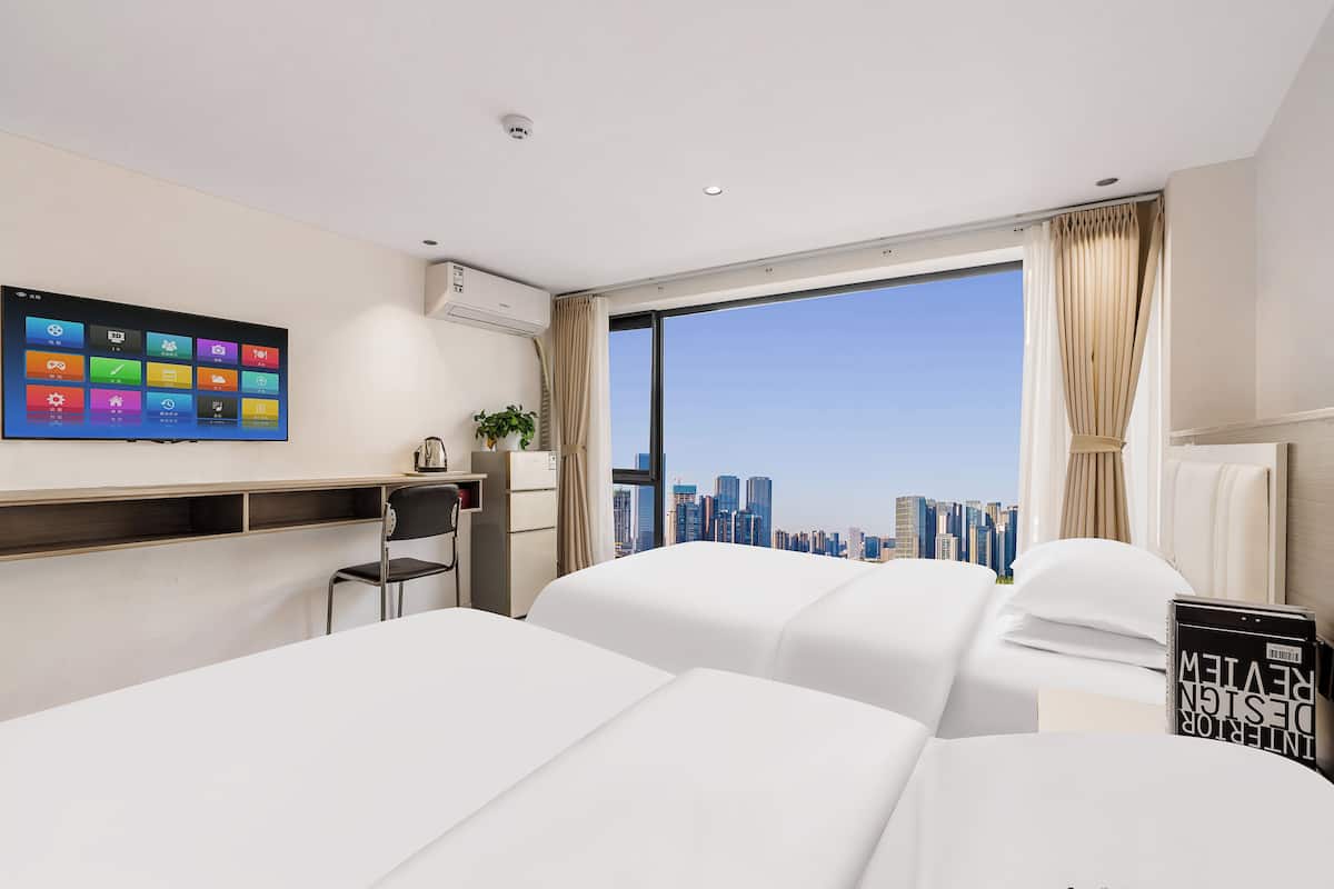 A modern hotel room with a large window, a flat-screen TV, a desk, and two beds.