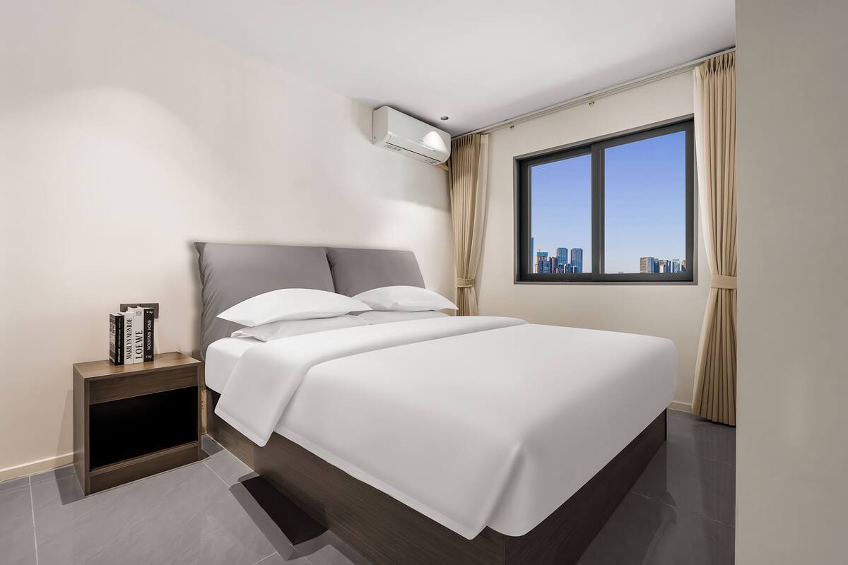 A modern bedroom with a large bed, a nightstand, an air conditioning unit, and a window with a city view.