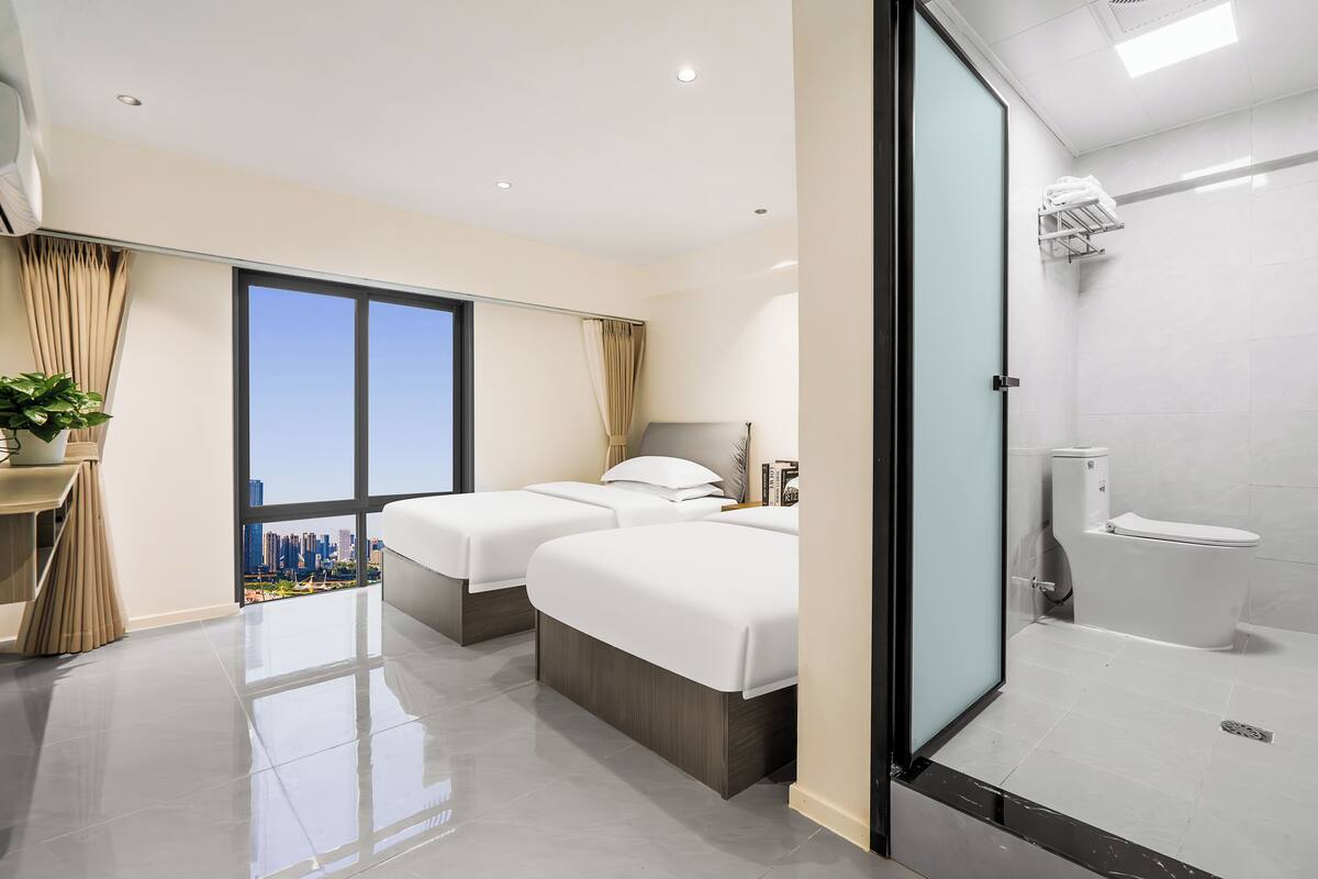 A modern hotel room with two beds, a large window with a city view, a bathroom with a toilet and shower, and a small plant on a side table.