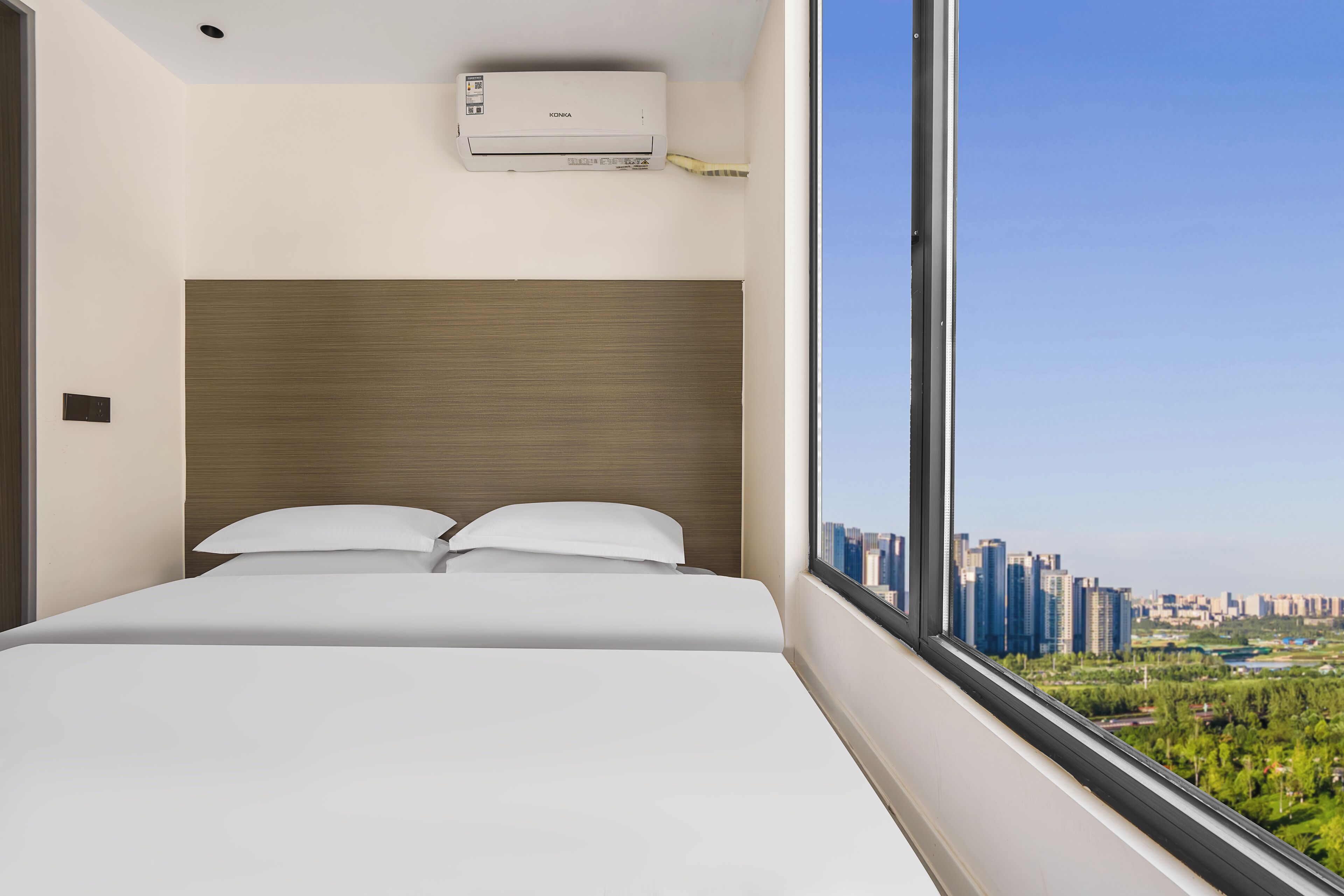 Une chambre d’hôtel moderne dotée d’un grand lit, d’une tête de lit en bois, d’une fenêtre donnant sur la ville et d’un appareil de climatisation.