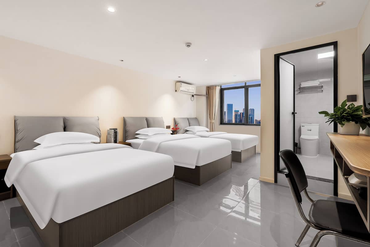 A modern hotel room with three beds, a desk, and a view of the city skyline.