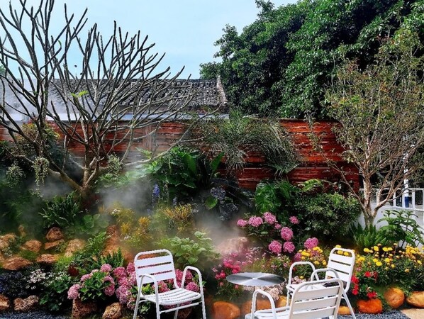 Ein Garten mit einer Backsteinmauer, verschiedenen Pflanzen und zwei weißen Metallstühlen um einen kleinen Tisch.