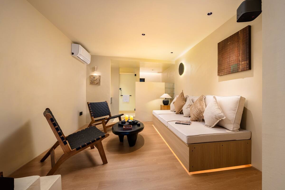 A modern living room with a wooden floor, a black chair, a round coffee table, a sofa with cushions, and a wall-mounted air conditioner.