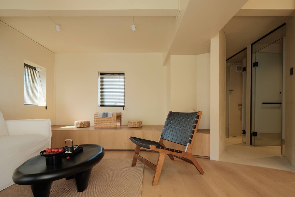 A modern living room with a black coffee table, a woven chair, and a sofa. There are two windows with blinds, a glass door, and a light-colored wall.
