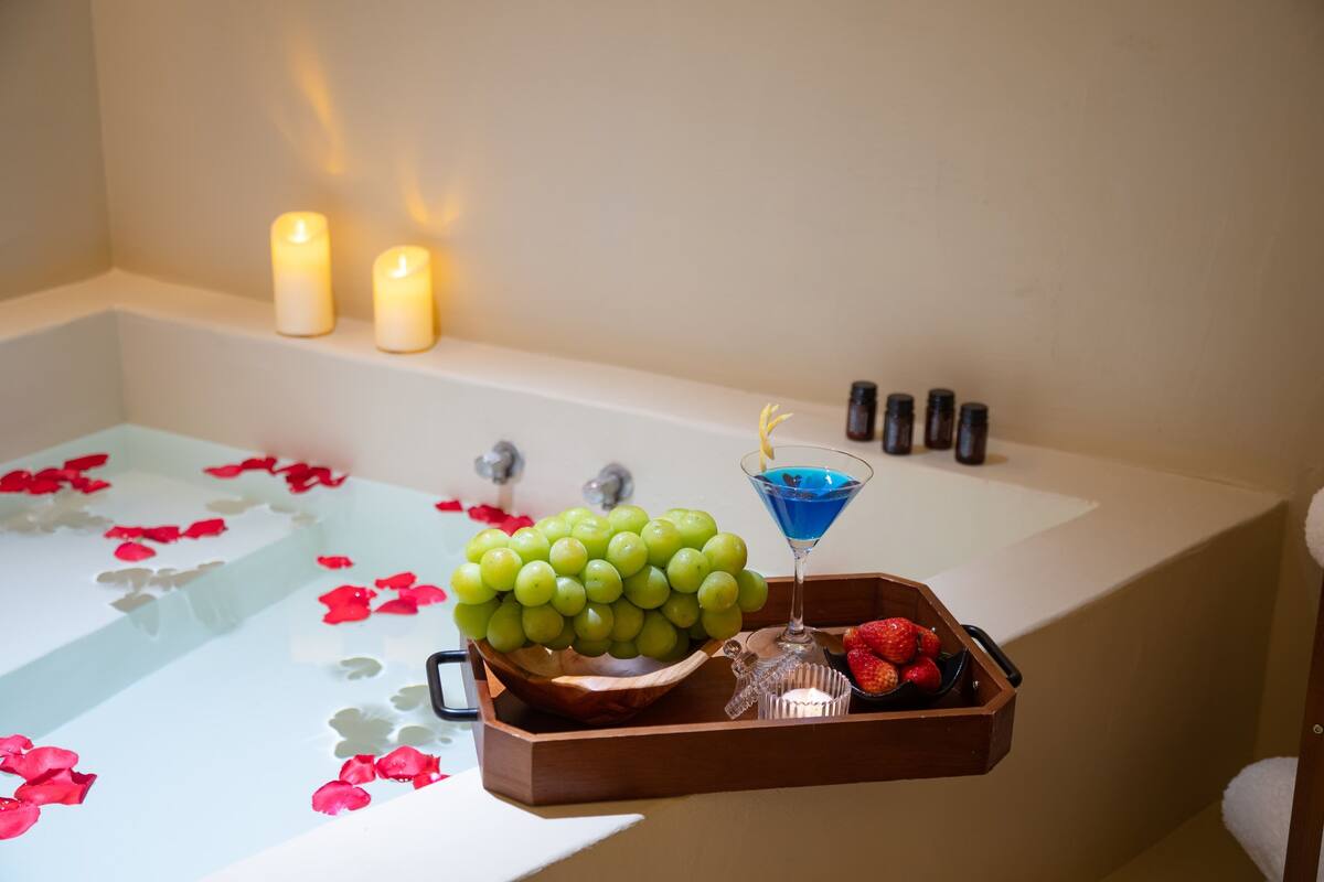 A bathtub with rose petals, a wooden tray with grapes, a martini glass, and a small bowl of strawberries.