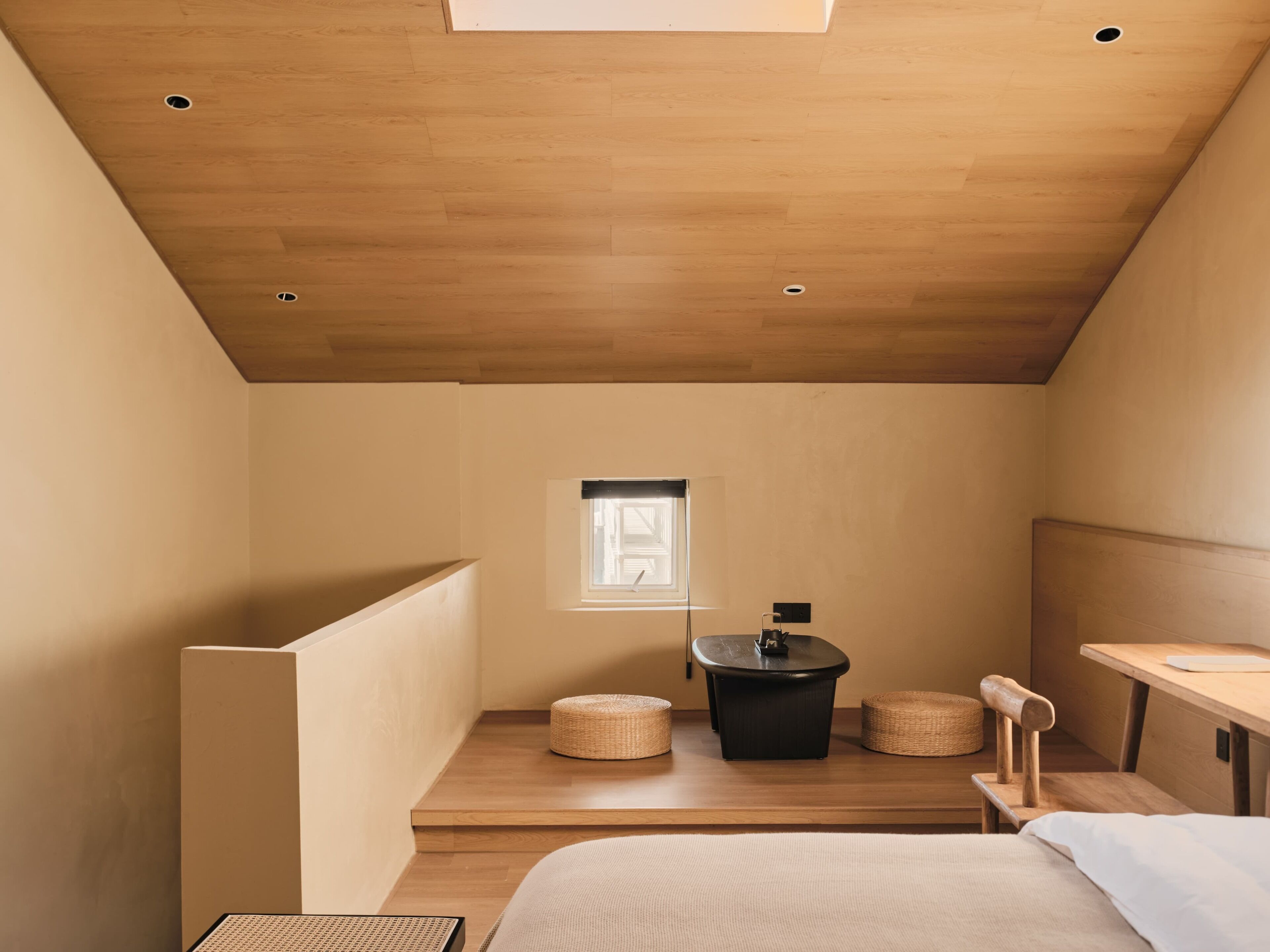 A compact, modern bedroom with a sloped ceiling, a small window, a round black table, and a wooden bench.