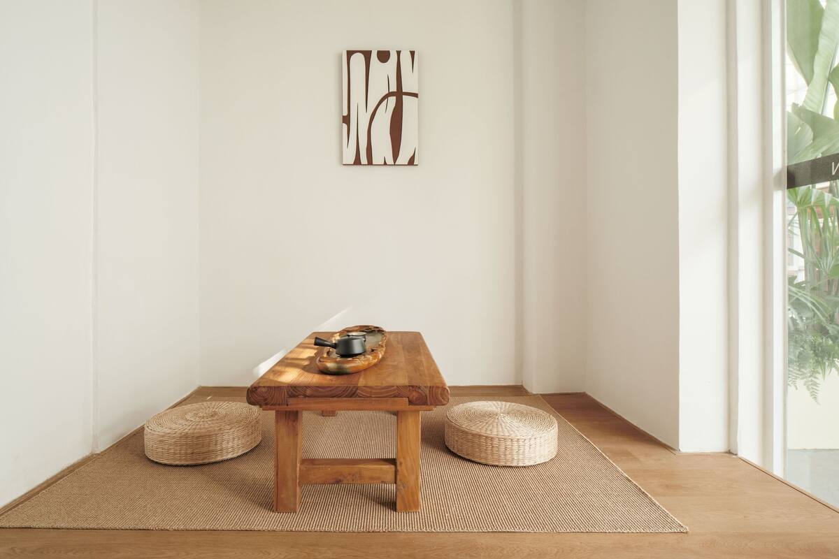 A minimalist room with a wooden table, woven poufs, and a wall-mounted abstract art piece.