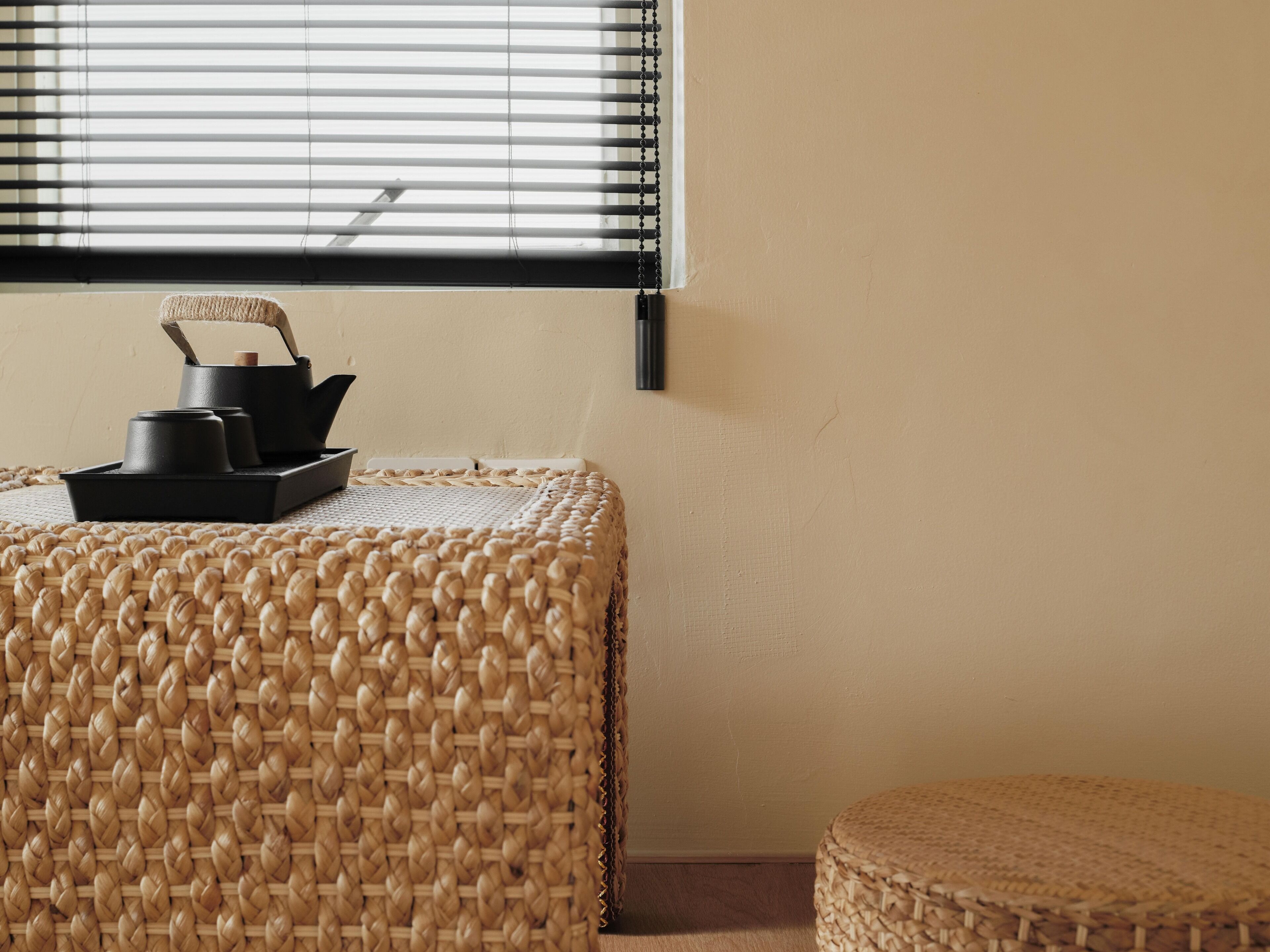 A corner of a room with a woven pouf, a black tray with items, and a window with blinds.