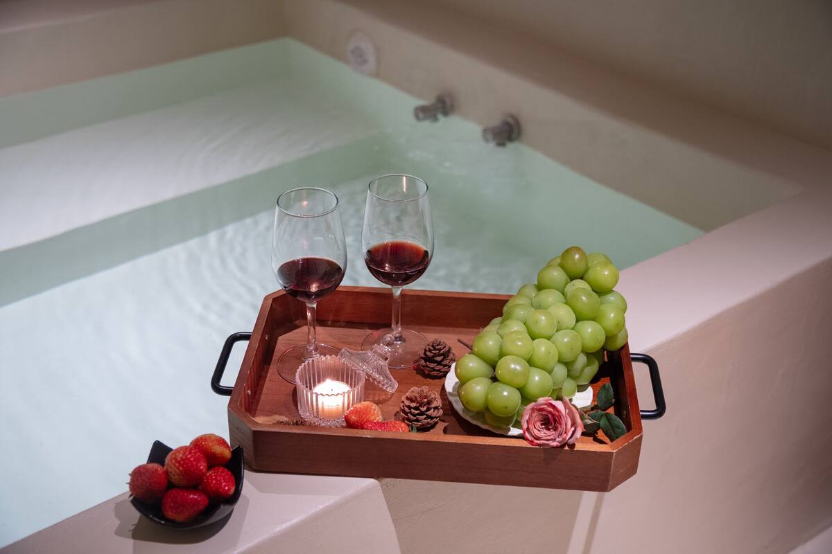 A tray with two glasses of red wine, a bowl of strawberries, a candle, a pine cone, a bunch of green grapes, and a rose.