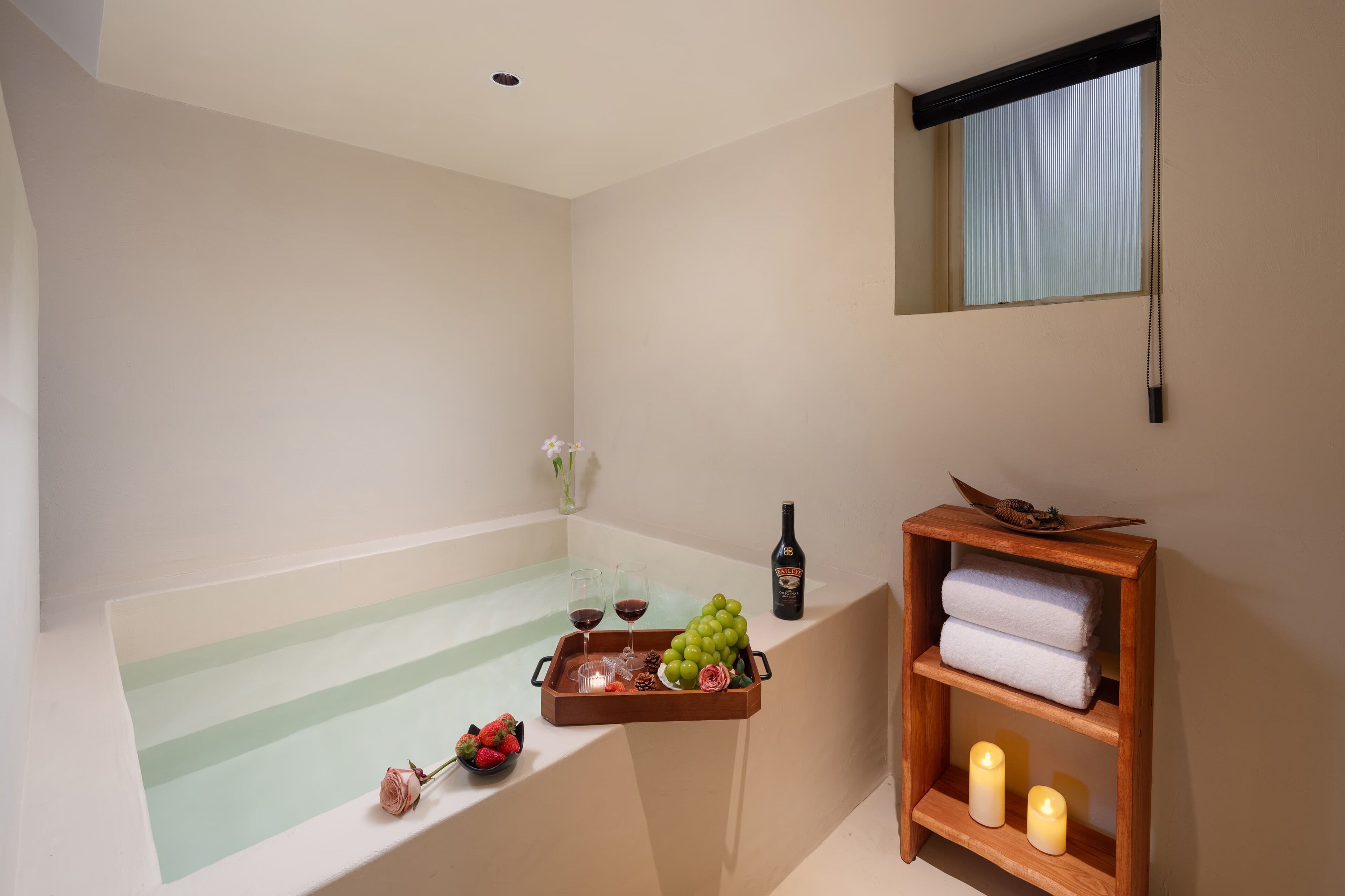 A bathroom with a bathtub, a wooden shelf with towels, a tray with grapes and glasses, and a bottle of wine.