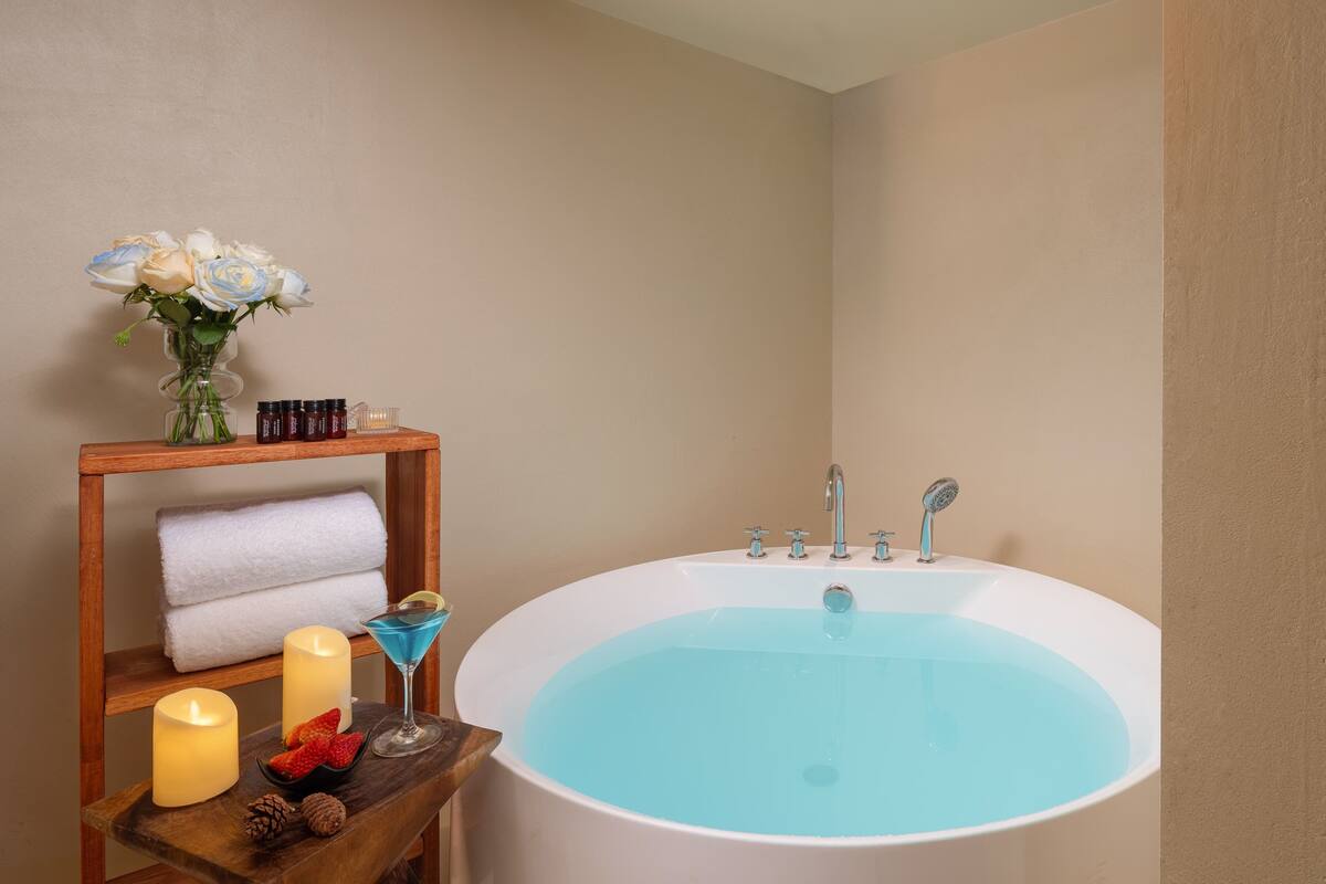 A bathroom with a large bathtub, a shelf with candles, a vase with flowers, and a tray with a drink and snacks.