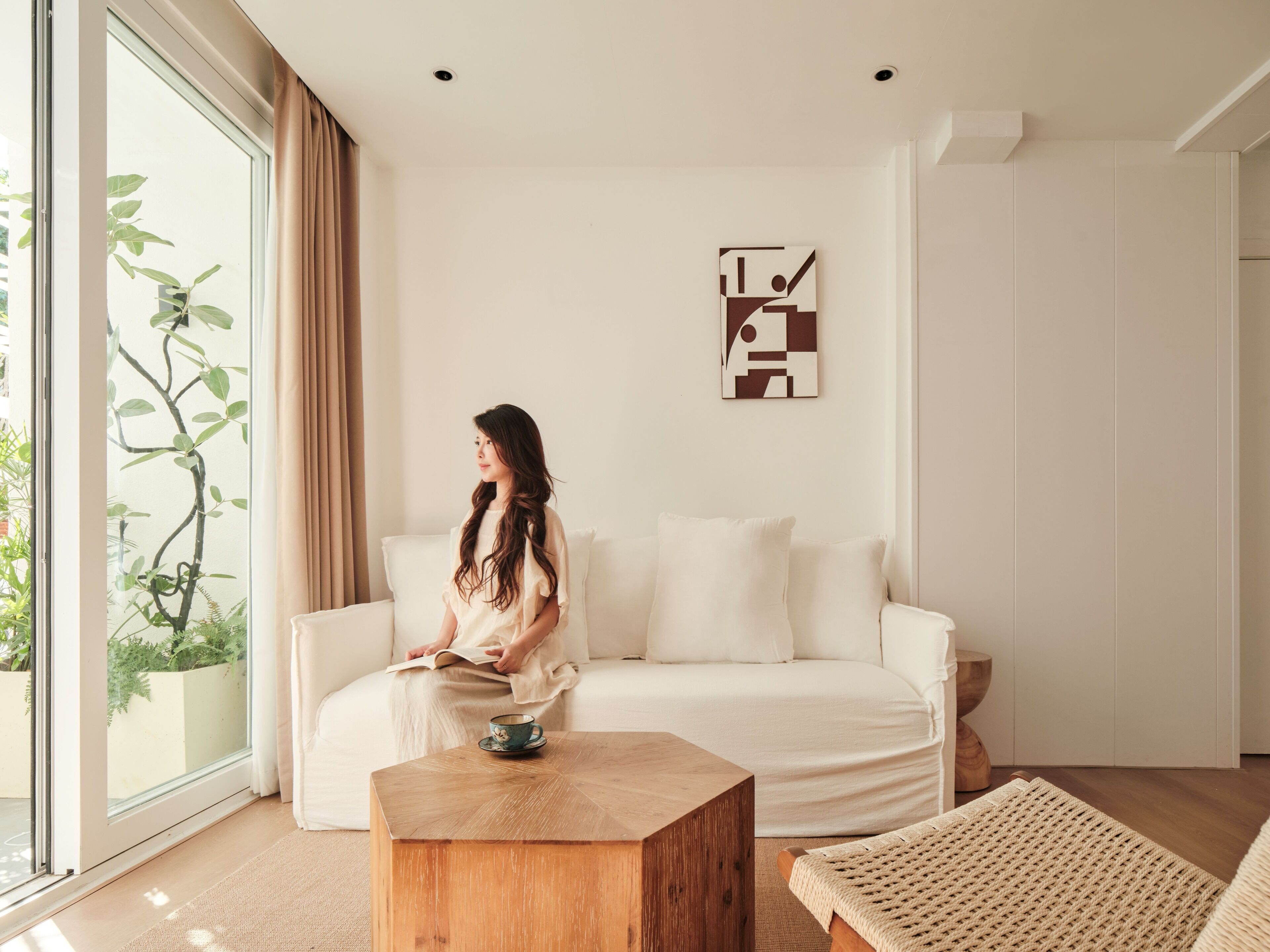 Ein modernes Wohnzimmer mit einem weißen Sofa, einem Holztisch und einem Gewebesessel. Eine Person sitzt auf dem Sofa, und ein Gemälde hängt an der Wand.