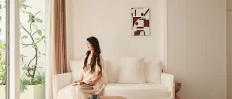 A modern living room with a white sofa, a wooden coffee table, and a woven chair. A person is seated on the sofa, and a painting is on the wall.