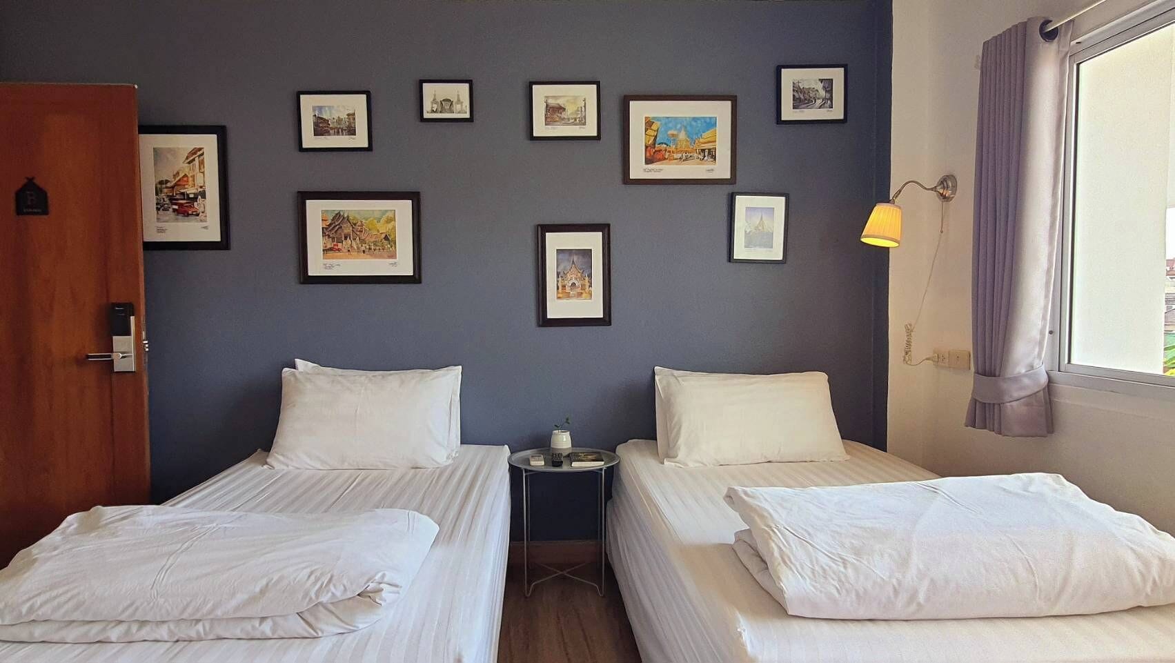 A hotel room with two beds, a small table, a lamp, and a wall decorated with framed pictures.