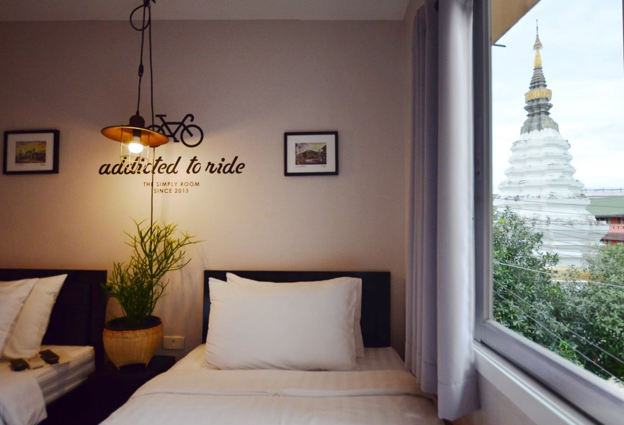 Una habitació d'hotel amb un llit, una planta en test, una làmpada fixa a la paret i una finestra amb vista a una pagoda blanca.