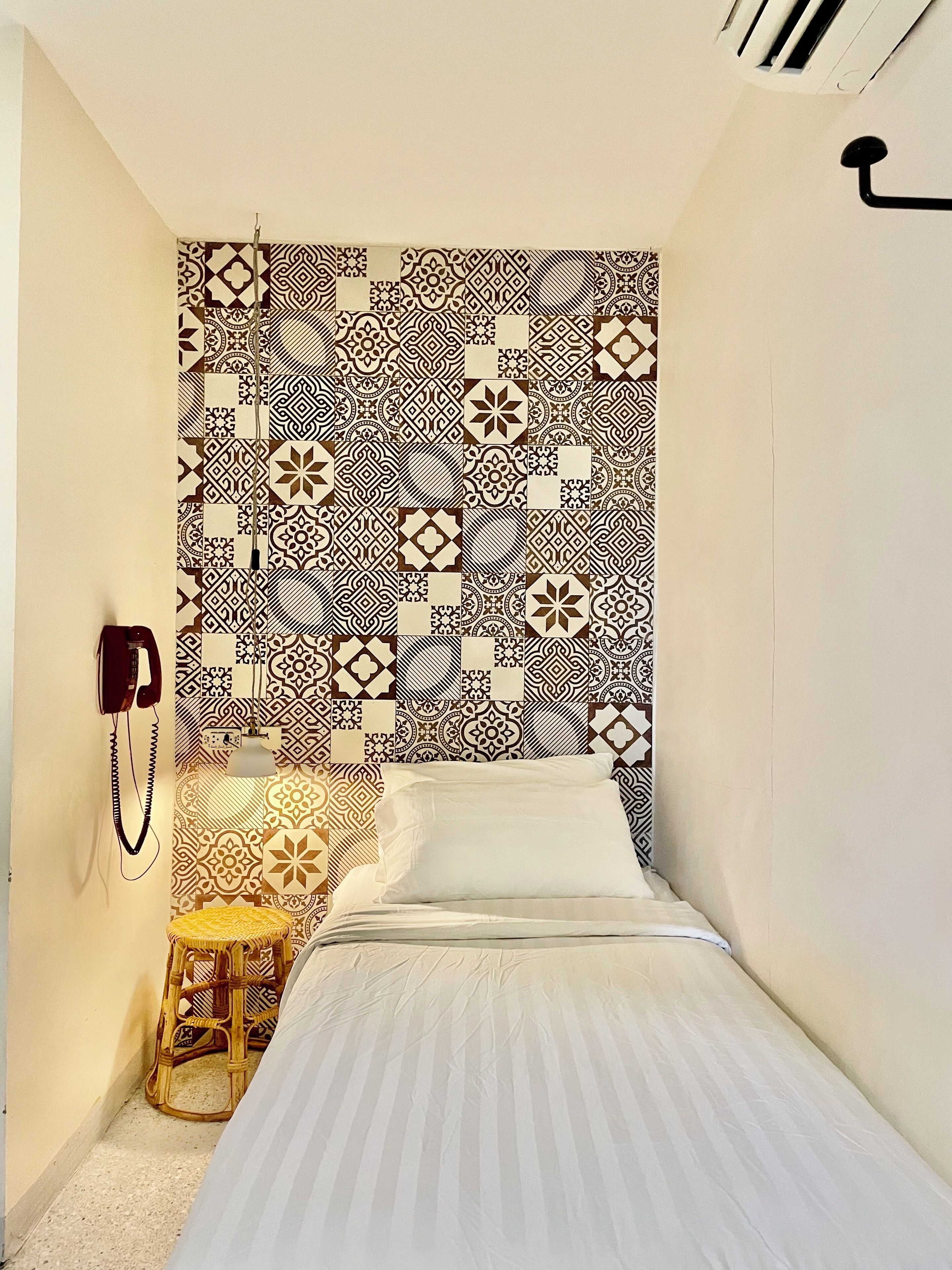 Une petite pièce étroite avec un mur orné de motifs, un lit, un petit tabouret en bois et un téléphone fixé au mur.