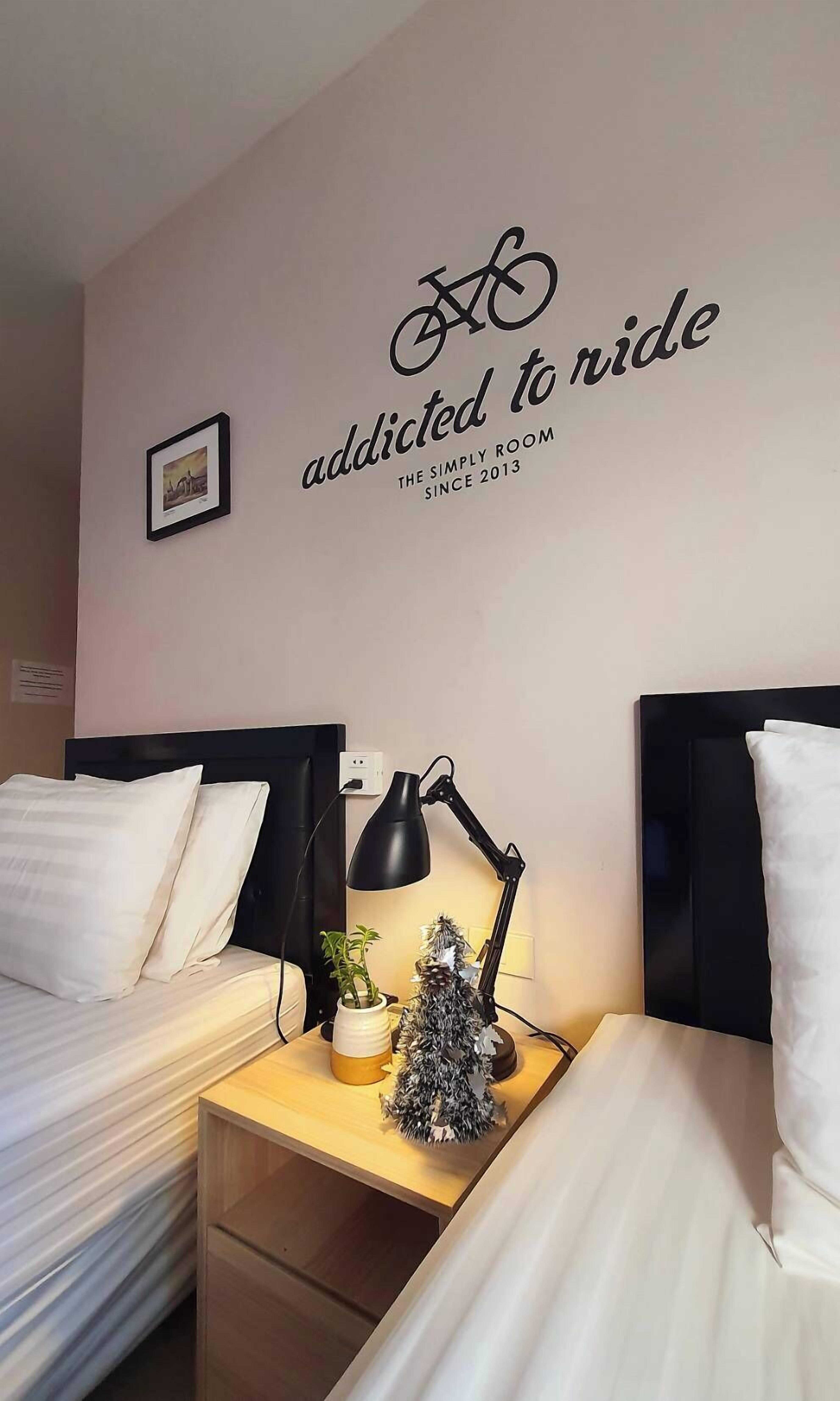 A neatly arranged hotel room with two beds, a wall-mounted sign reading "addicted to ride", and a small framed picture.