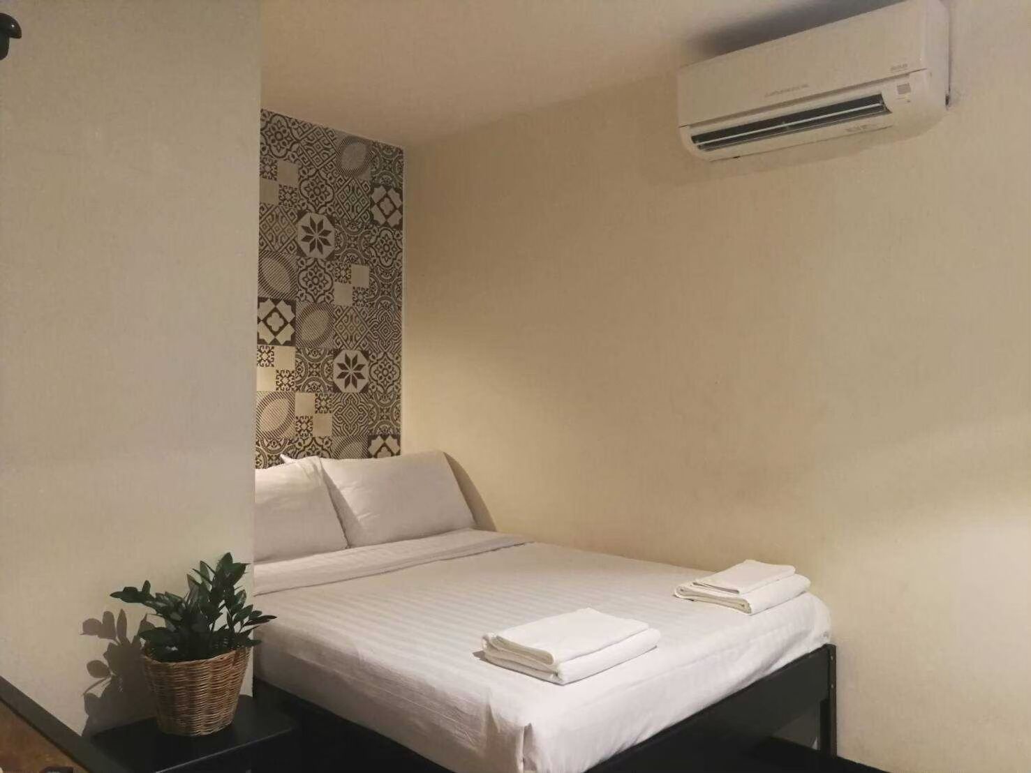 A small, neatly made bed with white linens and two folded towels, a potted plant on a nightstand, and a wall-mounted air conditioner.