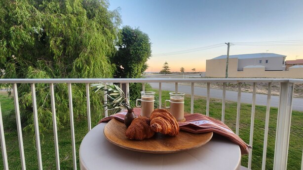 Ein Balkon mit Tisch, gedeckt für zwei Personen, mit Croissants und zwei Gläsern eines braunen Getränks, mit Blick auf ein Wohngebiet mit Häusern und blauem Himmel.