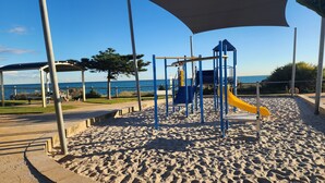 Ein Spielplatz mit Rutsche und Klettergerüsten, auf sandigem Untergrund mit Blick aufs Meer.
