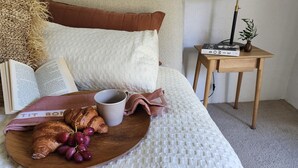 Ein ordentlich bezogenes Bett mit weißer Strukturware, ein Nachttisch aus Holz mit Lampe, Büchern und einer Auflage mit Croissants und Trauben.