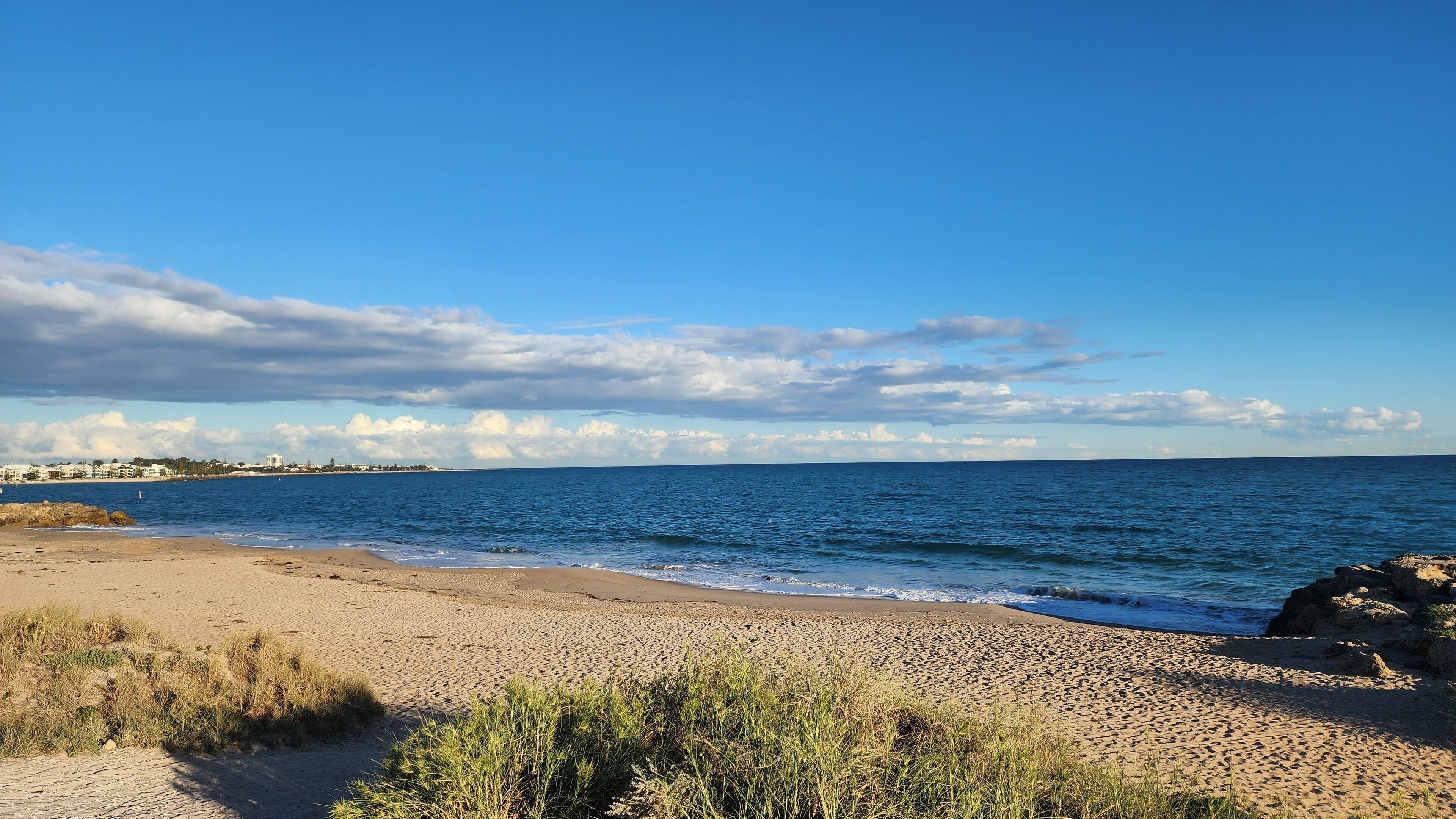 Une plage avec un ciel dégagé, une mer calme et des bâtiments qui se dessinent à l’horizon.