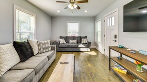 Un salon avec un canapé gris, une table basse en bois, un ventilateur de plafond et un téléviseur fixé au mur.
