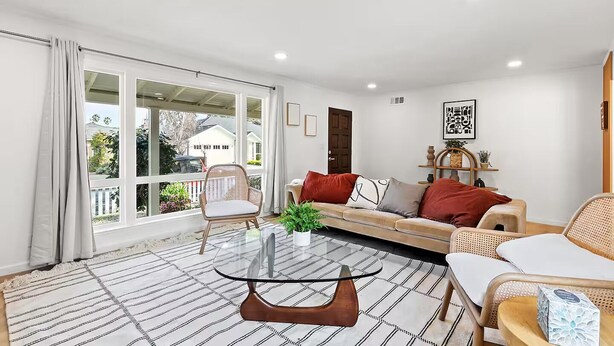 A modern living room with a glass coffee table, a sofa, and a chair. There is a large window with curtains, a door, and a small plant on the windowsill. - 222 - New, Stylish Quiet 2B1B w/ Yard near Stanford (Redwood City)