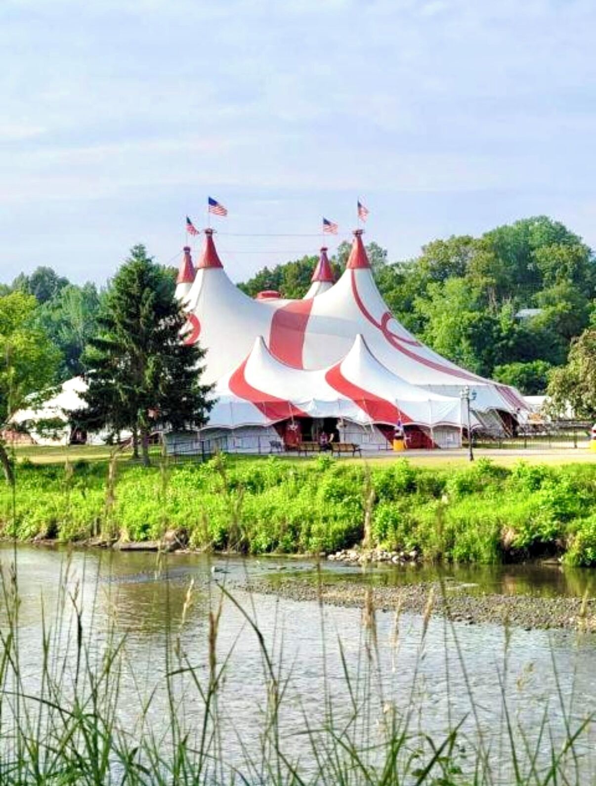 A large, white and red tented structure with multiple flags, located near a body of water and surrounded by greenery.