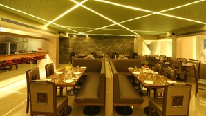 A restaurant with a modern interior, featuring long communal tables and booths, a kitchen area visible to the left, and a stone wall in the background. - The Maharaja Business Hotel (Mumbai)