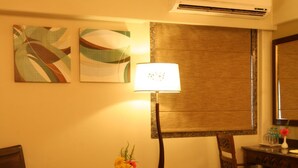A hotel room with a bed, a desk with a lamp, two chairs, a small table with a fruit basket and flowers, and abstract wall art. - The Maharaja Business Hotel (Mumbai)