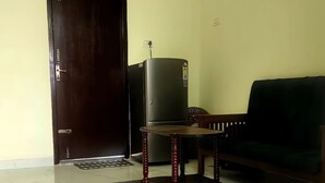 A room with a dark wooden door, a black refrigerator, a wooden table with a black mat, and a dark sofa. - Sai Home Stay (Coimbatore)