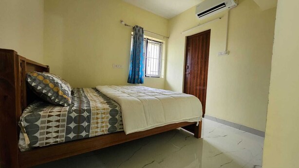 A bedroom with a bed, a window with curtains, an air conditioning unit, and a wooden door. - Sai Home Stay (Coimbatore)