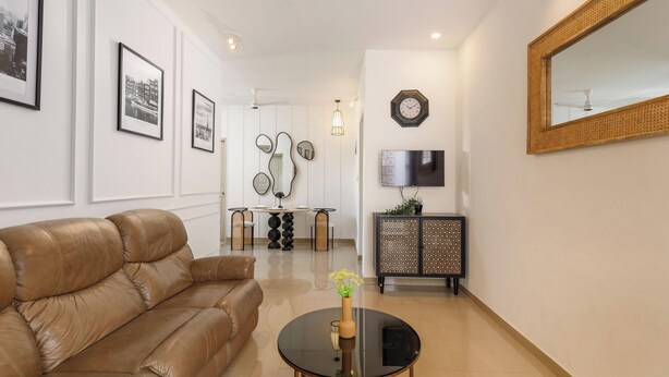 A modern living room with a brown leather sofa, a round glass coffee table with a vase, a dining area with a round table and chairs, and a TV on the wall. - Golden Ivory by Red Olive (Bengaluru)