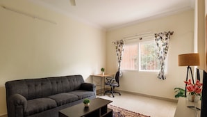 A living room with a dark grey sofa, a small wooden table with a potted plant, a black office chair, a TV on a stand, and a window with floral curtains. - The City Nova Apartment Koramangla (Bengaluru)