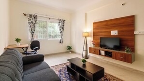 A living room with a sofa, a coffee table, a TV on a wooden cabinet, a lamp, and a window with curtains. - The City Nova Apartment Koramangla (Bengaluru)