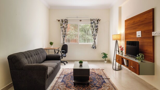 A living room with a sofa, a coffee table, a TV on a wooden cabinet, a small table with a lamp, and a window with curtains. - The City Nova Apartment Koramangla (Bengaluru)